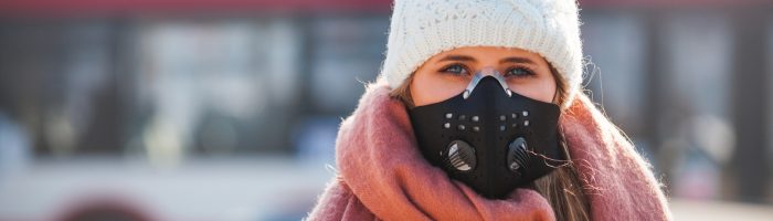 Jak chronić się przed smogiem w Polsce? Jaką maskę antysmogową wybrać? Kobieta w białej czapce, różowym szaliku i masce antysmogowej stoi zimą na ulicy w mieście.