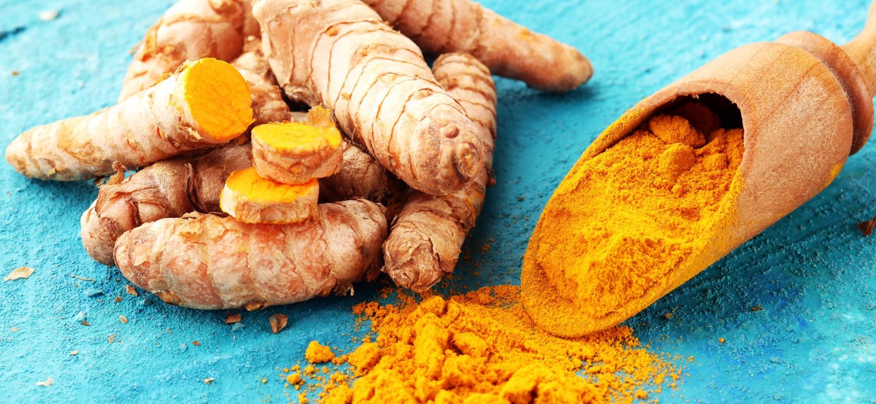 Napój z kurkumy - jak go przygotować? Korzeń kurkumy i sproszkowana kurkuma na drewnianej łyżce leżą na niebieskim blacie.