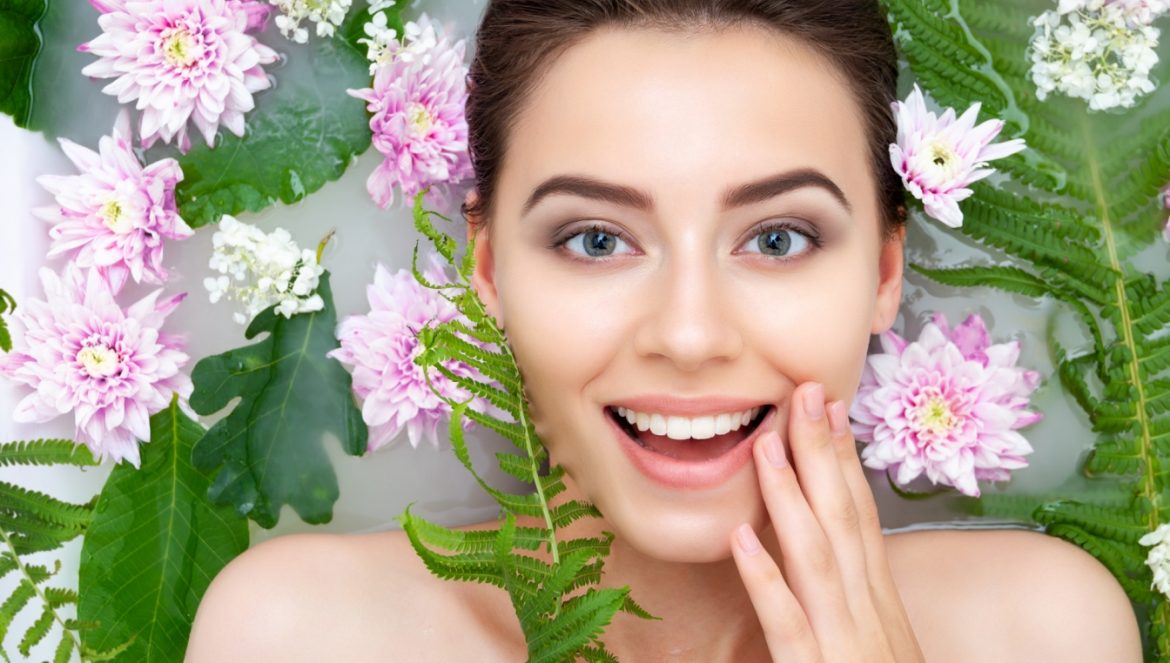 6 kwiatów, które pokocha twoja skóra! Sprawdź, jakie wyciągi z kwiatów najlepiej pielęgnują skórę. Portret kobiety, która bierze kąpiel wśród kwiatów i liści paproci.