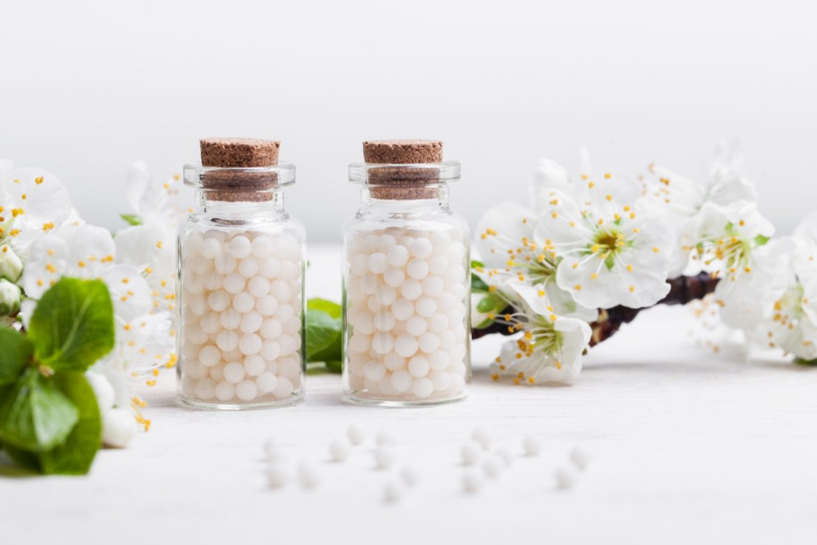 Sepia officinalis - lek homeopatyczny na różne dolegliwości - fizyczne i emocjonalne. Dwie fiolki granulek homeopatycznych stoją na białym blacie w otoczeniu białych kwiatów jabłoni.