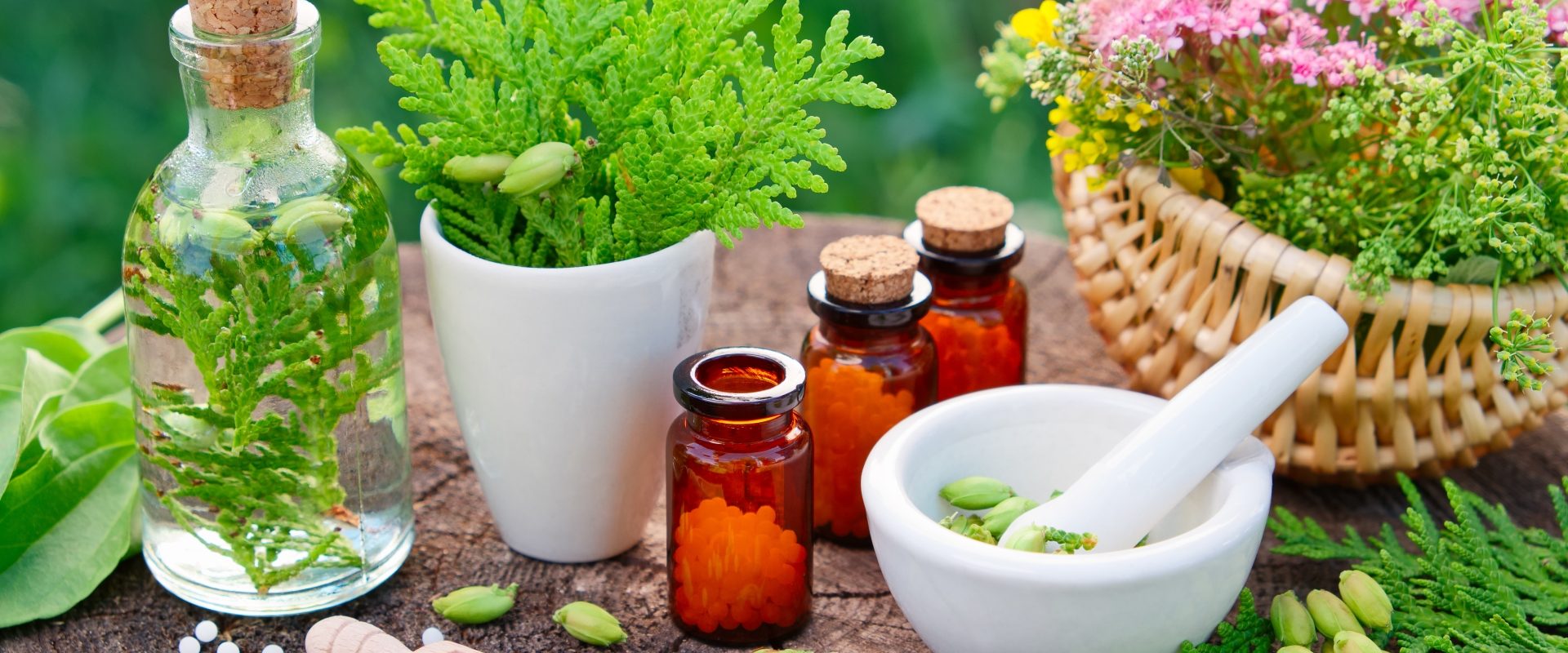 Naturalne sposoby na wirusy - leki roślinne i leki homeopatyczne. Fiolki z lekami homeopatycznymi i zielone rośliny oraz kwiaty w koszyku i wazonikach stoją na drewnianym stole w ogrodzie.