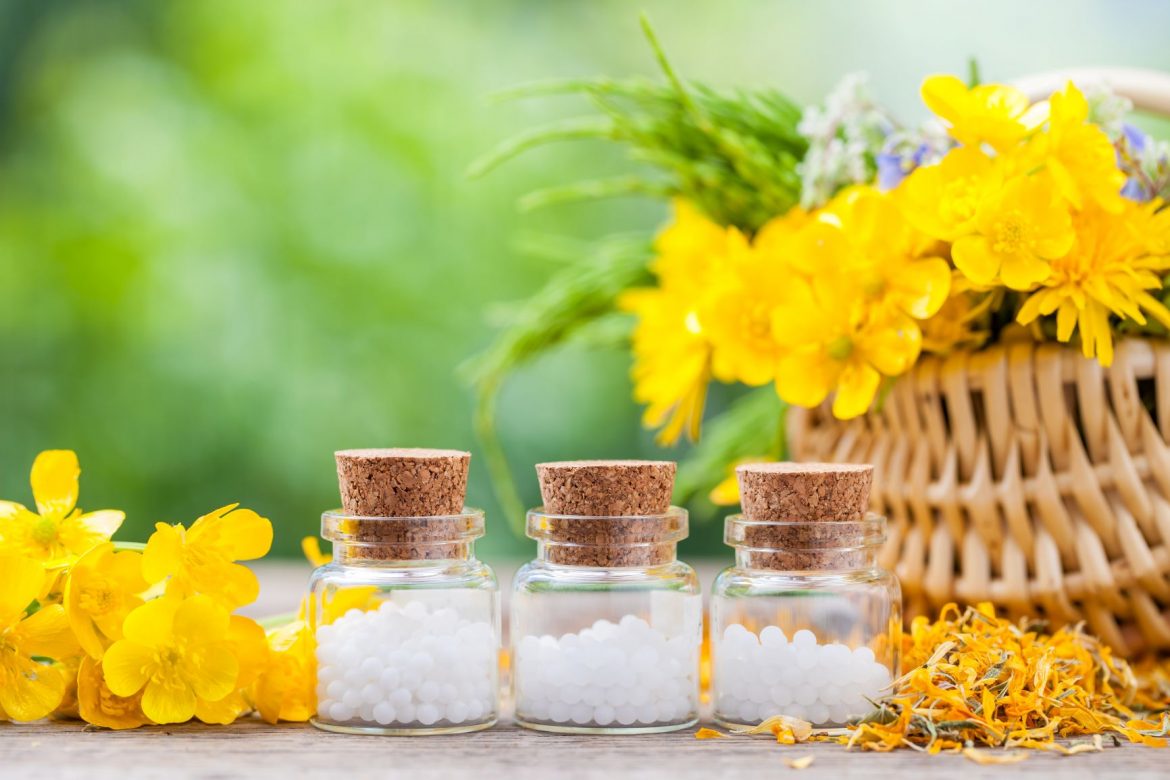 Naturalny sposób na brodawki. Jakie zioła i rośliny lecznicze pomagają na brodawki? Jakie leki homeopatyczne na brodawki stosować? Granulki homeopatyczne w trzech fiolkach stoją na stole w ogrodzie obok kosza wiklinowego z żółtymi kwiatami.