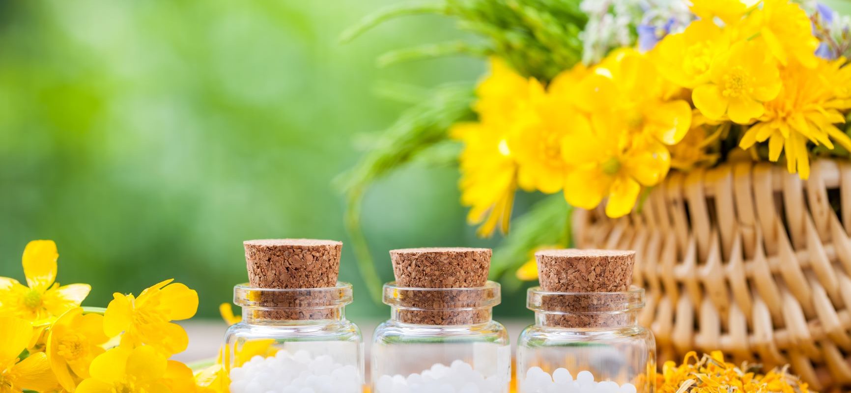Naturalny sposób na brodawki. Jakie zioła i rośliny lecznicze pomagają na brodawki? Jakie leki homeopatyczne na brodawki stosować? Granulki homeopatyczne w trzech fiolkach stoją na stole w ogrodzie obok kosza wiklinowego z żółtymi kwiatami.