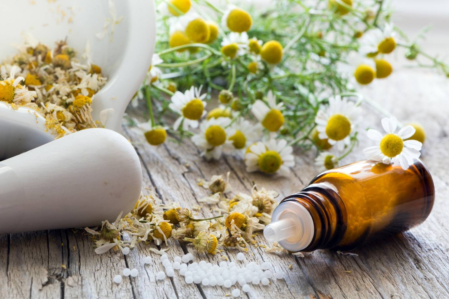 Leczenie traumy homeopatią. Jakie leki homeopatyczne pomogą poradzić sobie ze stresem traumatycznym? Fiolka leków leży obok białego moździerza i kwiatów rumianku.