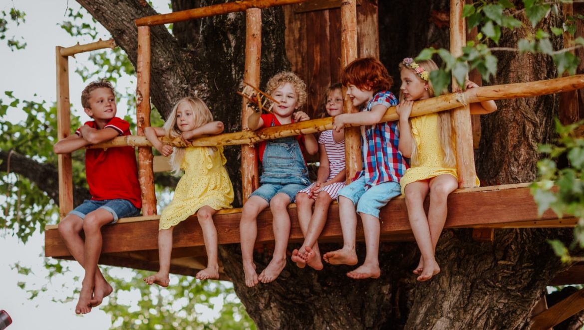Leśne przedszkole - czym jest, jakie są jego zalety, dlaczego warto posłać do niego dzieci? Przedszkolaki starszaki siedzą w domku na drzewie w lesie.