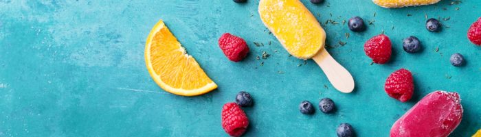 Przepisy na domowe lody naturalne. Owocowe lody na patyku leżą na niebieskim tle, obok rozrzucone owoce.