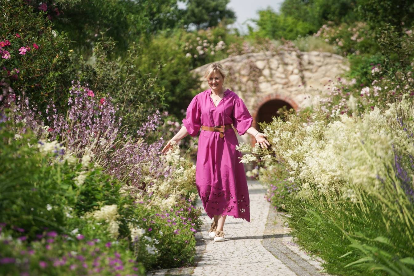 Maja Popielarska (Maja w ogrodzie) spaceruje po ogrodzie w fioletowej sukience.