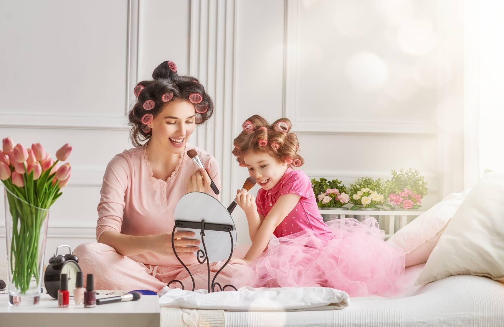 Relacje matka - córka. Mama i córeczka siedzą po turecku na podłodze w wałkach na włosach i malują się przed lusterkiem.