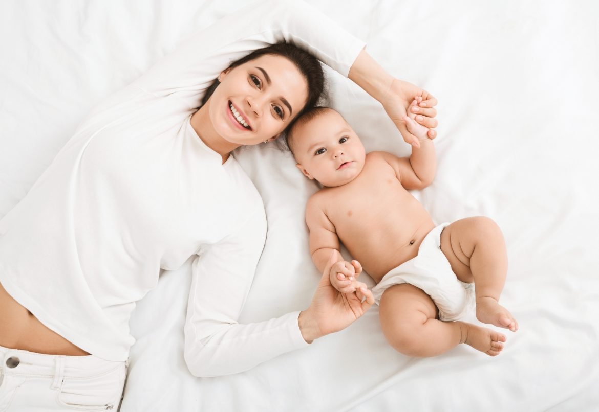 Calcarea carbonica dla dzieci i dorosłych. Szczęśliwa mama w białej bluzce leży z niemowlęciem w pieluszce na łóżku i trzyma go za rączki.