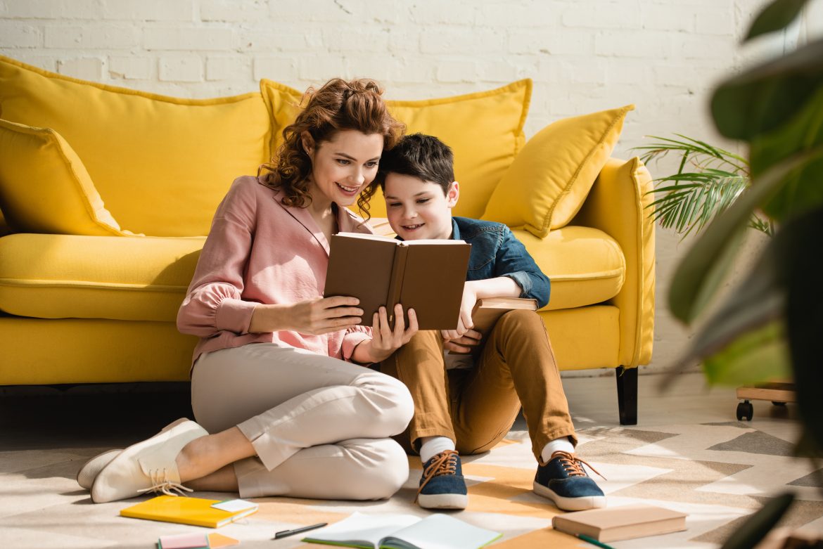 Jak wychować syna w szacunku do kobiet? Mama i syn w wieku szkolnym wspólnie czytają książkę w salonie, opierając się o żółtą kanapę.