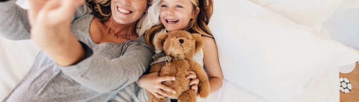 Matka i córka - jak zbudować i ułożyć sobie dobrą relację? Mama i córka przytulająca misia leżą na łóżku i robią sobie selfie.
