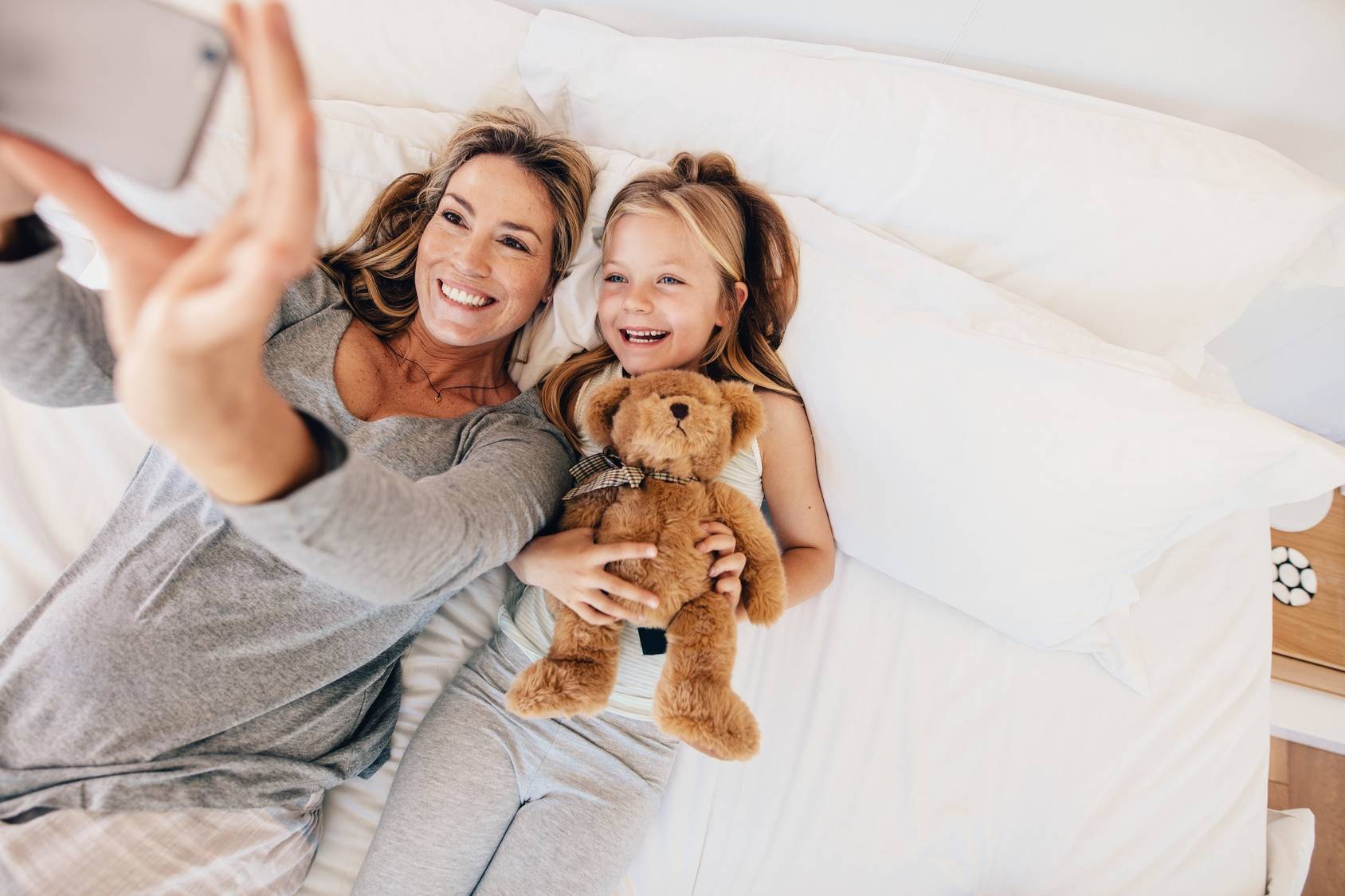 Matka i córka - jak zbudować i ułożyć sobie dobrą relację? Mama i córka przytulająca misia leżą na łóżku i robią sobie selfie.