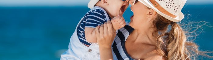 Udar słoneczny u dzieci, poparzenie słoneczne. Mama trzyma uśmiechnięte niemowlę na rękach na plaży.