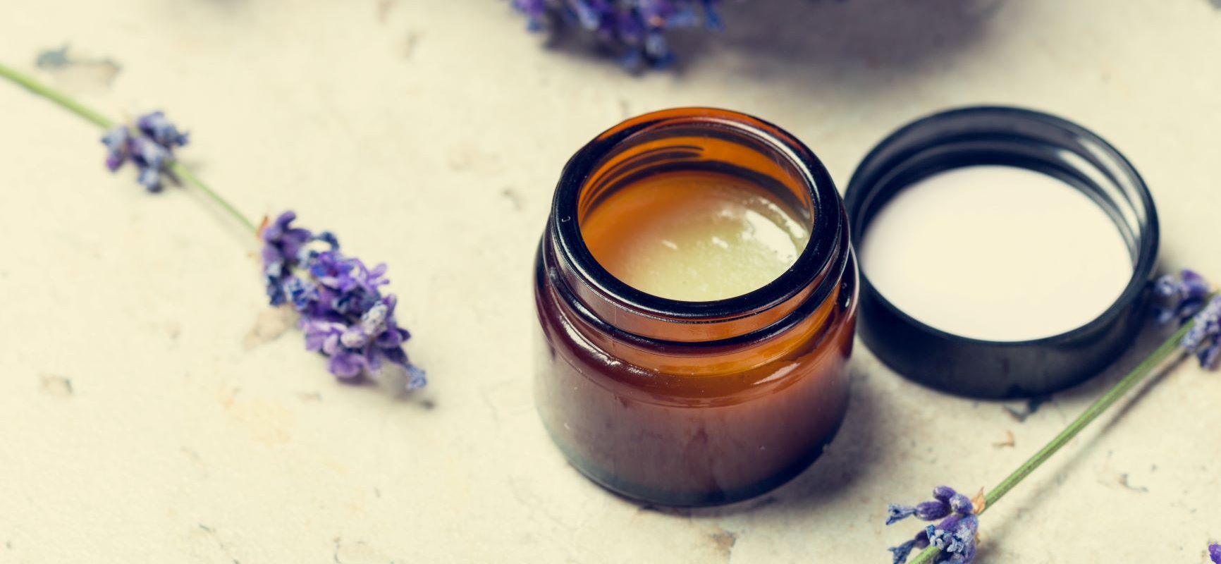 Maść rozgrzewająca na przeziębienie - jak ją zrobić? Otwarty słoiczek z maścią leży na blacie, obok niebieskie kwiaty.
