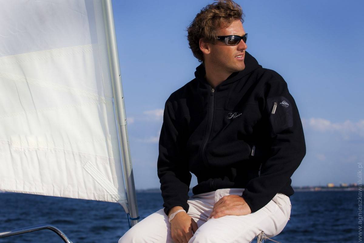 Mateusz Kusznierewicz, żeglarz i sportowiec, siedzi na łódce w czarnym polarze, białych spodniach i okularach przeciwsłonecznych.
