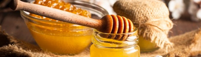 Propolis - co to jest kit pszczeli? Jakie właściwości ma propolis i jak go stosować. Na drewnianym rustykalnym stole stoi słoik miodu i miseczka z plastrem miodu, obok leżą białe kwiatki wiśni lub jabłoni.