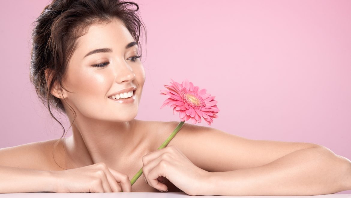 Jak zadbać o cerę naczynkową? Młoda kobieta o pięknej rumianej cerze na różowym tle opiera się o blat i trzyma w ręku różowy kwiatek.