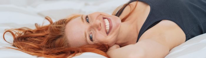 Jak wybrać najlepszy materac do spania? Rudowłosa kobieta leży na białym materacu w sypialni i uśmiecha się do aparatu.