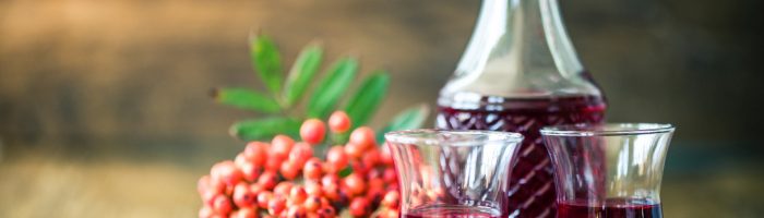 Przepisy na nalewki z jarzębiny, kasztanów i tarniny. Sprawdź, jakie mają właściwości lecznicze. Nalewka z jarzębiny w karafce i szklankach na drewnianym blacie.
