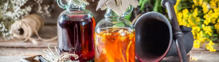 Przepisy na nalewki lecznicze. Na drewnianym blacie stoją dwie butelki z kolorowymi ziołowymi nalewkami. W tle wiszą zioła i leży moździerz.