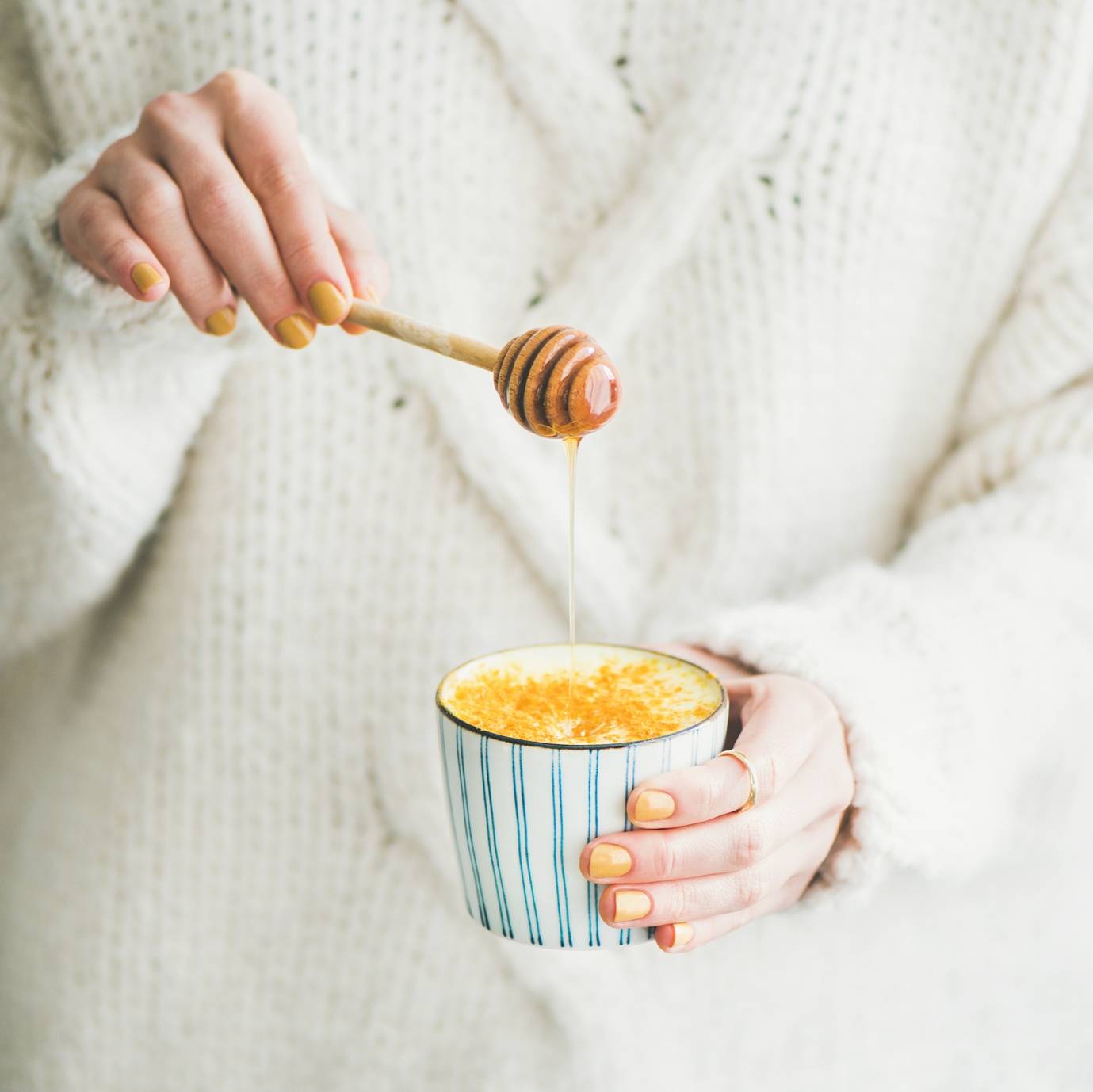 Jak w ciągu jednego dnia wyleczyć przeziębienie? Kobieta w ciepłym wełnianym swetrze trzyma kubek ciepłego mleka i drewnianą łyżkę do miodu. Zbliżenie na kubek i kobiece ręce z pomalowanymi na żółto paznokciami.