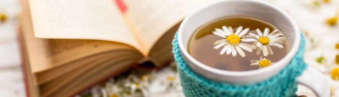 Jak leczyć przeziębienie według medycyny chińskiej? Kubek herbaty rumiankowej w sweterku stoi obok otwartej książki, wokół leżą kwiatki rumianku.