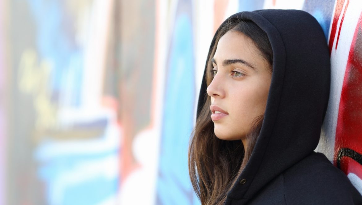 Nastolatki w pandemii - jak im pomóc? Nastoletnia skaterka w czarnej bluzie z kapturem opiera się o mur pomalowany graffiti.