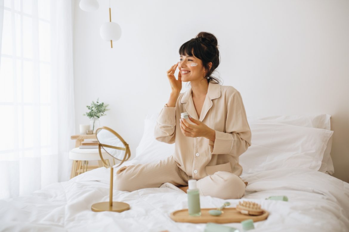 7 sposobów na naturalny demakijaż - jakie produkty roślinne pomagają dokładnie oczyścić twarz? Uśmiechnięta kobieta w beżowej piżamie siedzi na łóżku i pielęgnuje skórę twarzy.