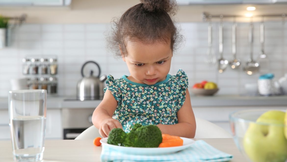 Neofobia żywieniowa - co robić, kiedy dziecko nie chce jeść? Mała dziewczynka podejrzliwie przygląda się warzywom na swoim talerzu.