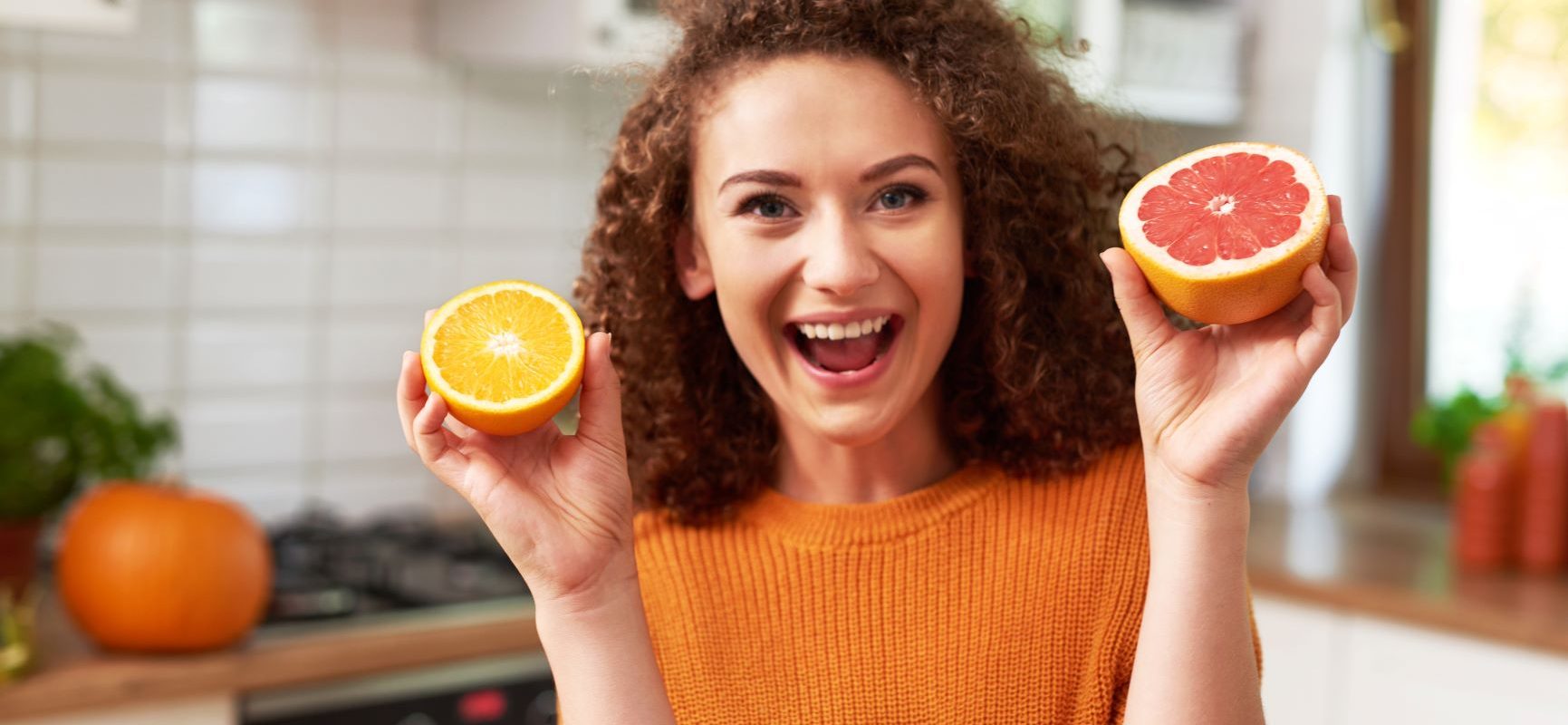 Niedobór witaminy C - jak rozpoznać objawy? Uśmiechnięta kobieta w kręconych włosach i pomarańczowym swetrze trzyma w rękach przepołowione cytrusy, za nią widać kuchnię.