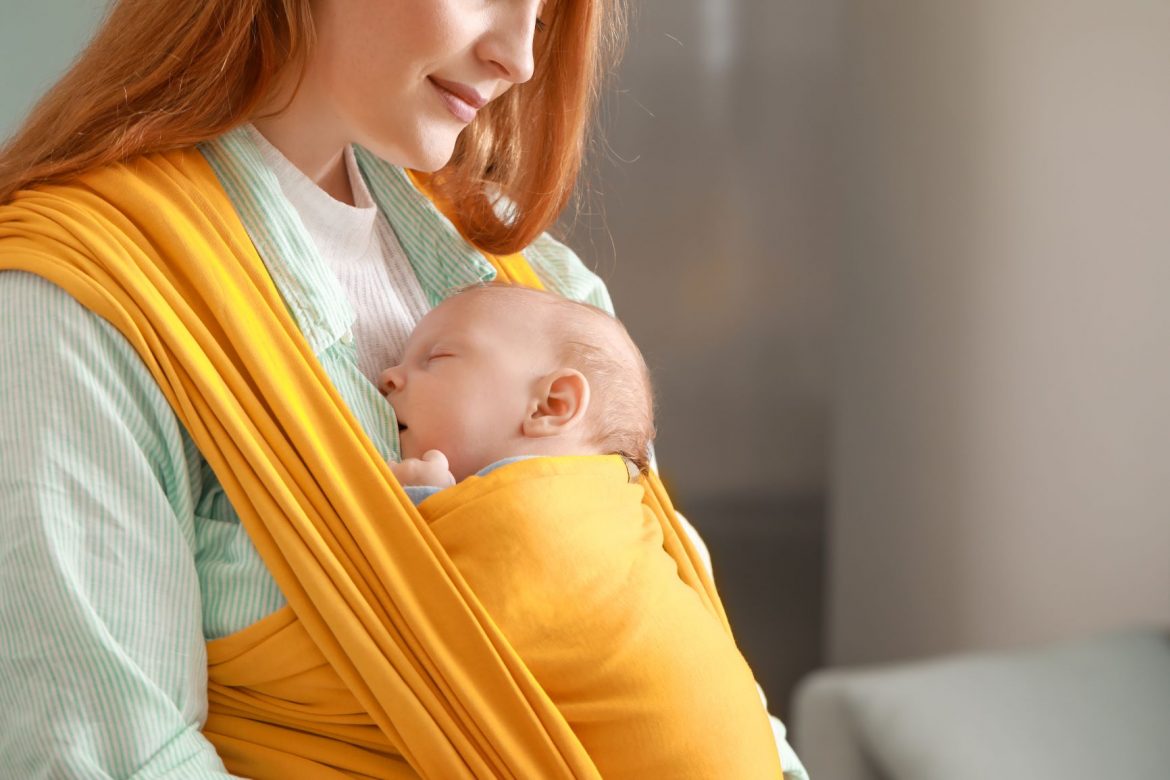 Chustonoszenie - jak nosić niemowlę w chuście? Mama tuli niemowlę zawinięte w żółtą chustę.