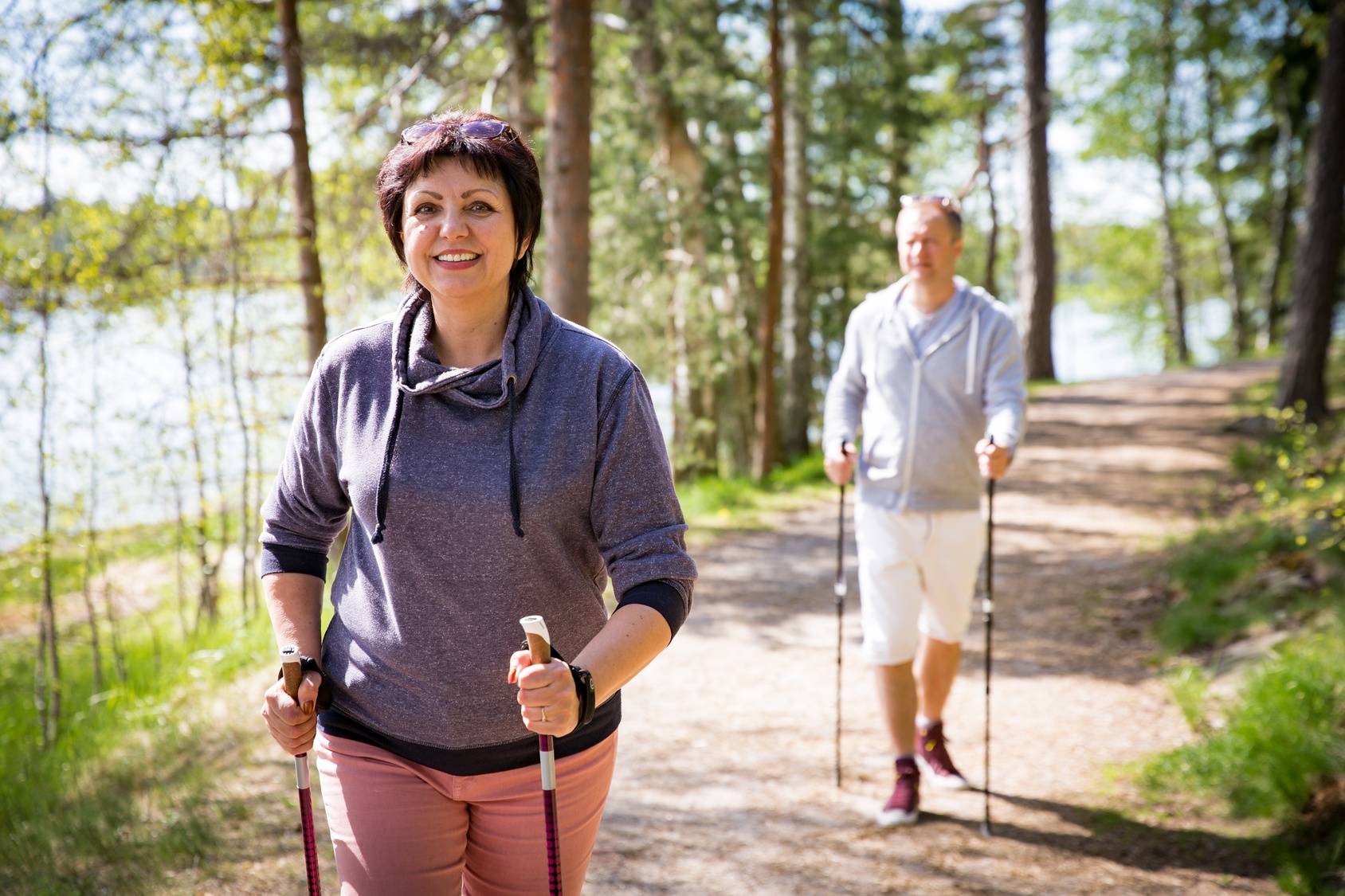 Nordic walking dla początkujących - gdzie i jak uprawiać ten sport? Mężczyzna i kobieta uprawiają nordic walking w lesie nad wodą.