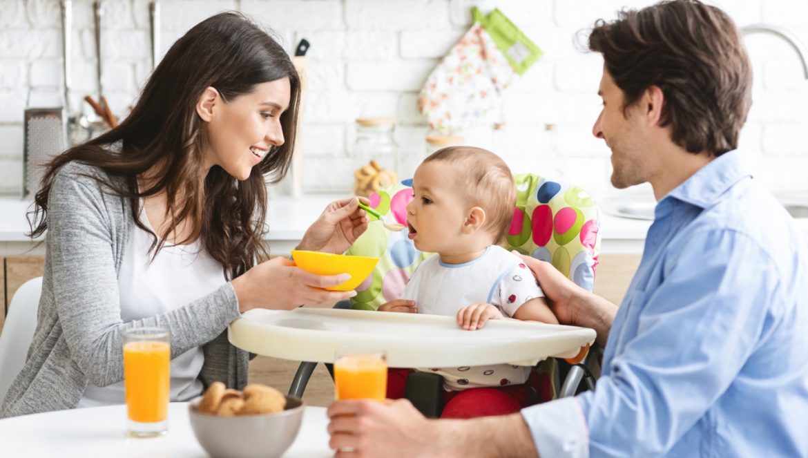 Nowy schemat żywienia niemowląt 2021 - jak karmić niemowlę i czym żywić dziecko? Mama i tata karmią niemowlaka siedzącego w swoim krzesełku przy stole w kuchni.