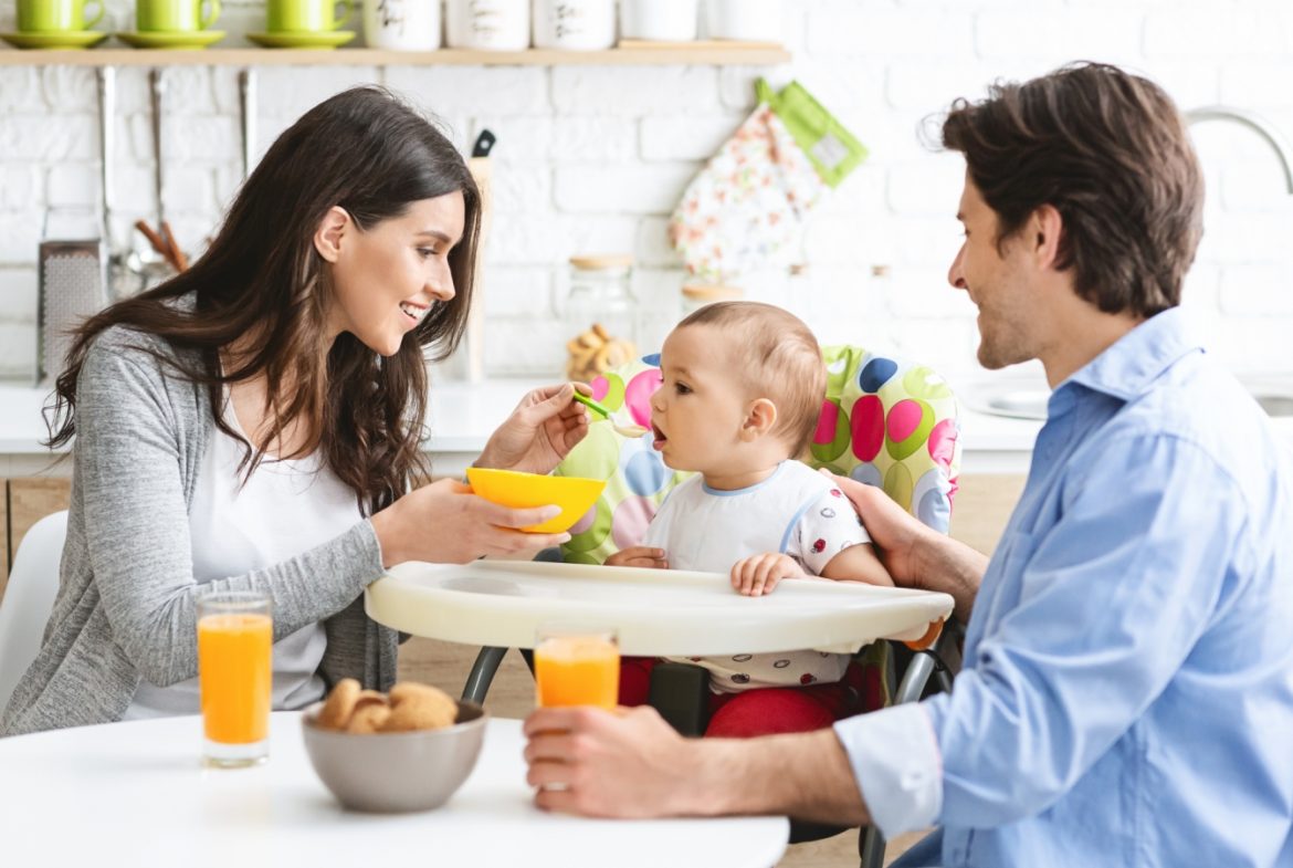 Nowy schemat żywienia niemowląt 2021 - jak karmić niemowlę i czym żywić dziecko? Mama i tata karmią niemowlaka siedzącego w swoim krzesełku przy stole w kuchni.