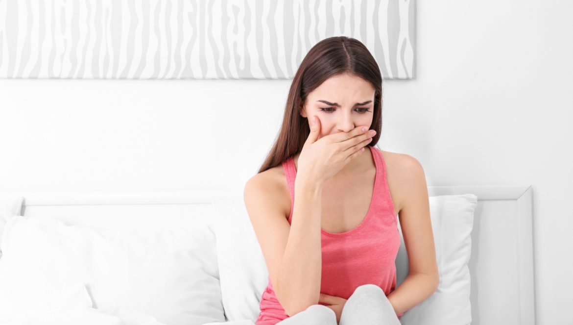Objawy ciąży - jak rozpoznać, czy doszło do zapłodnienia? Jakie są pierwsze objawy ciąży? Młoda kobieta w różowym topie i białych spodniach siedzi na łóżku i trzyma dłoń przy ustach z powodu mdłości.