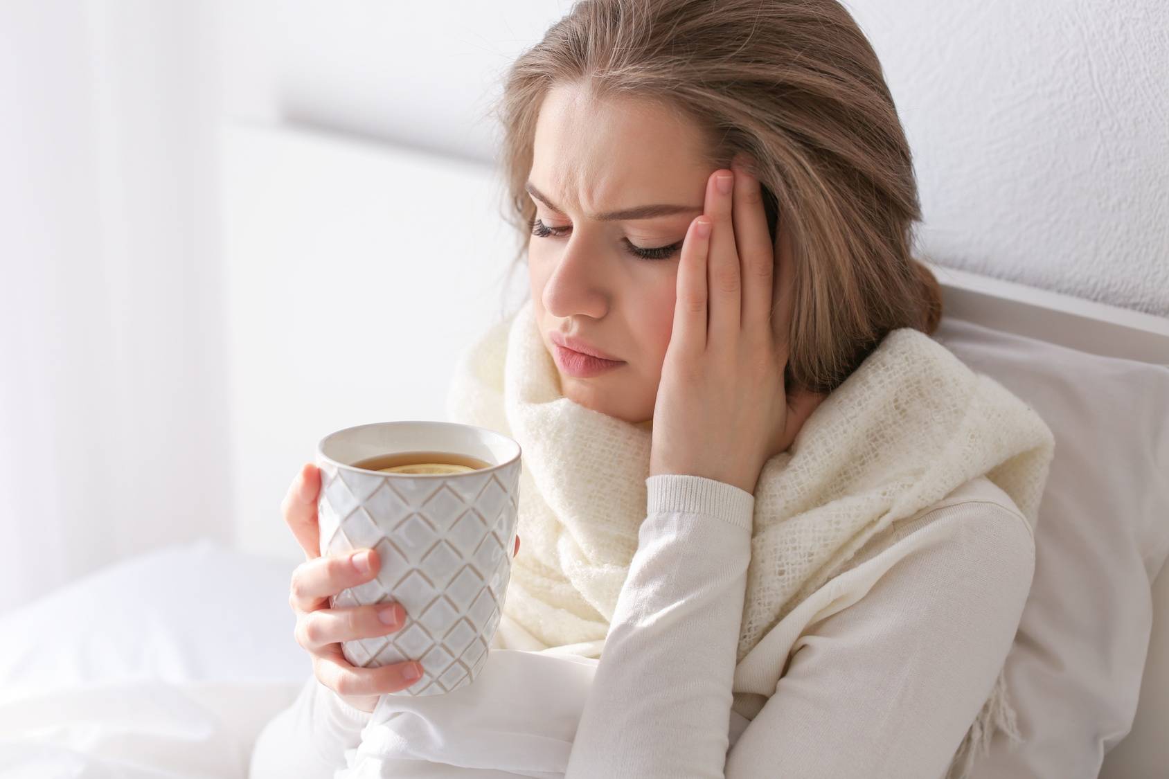 Objawy grypy - jak je rozpoznać? Kobieta leży w łóżku, dotyka skroni i pije gorącą herbatę z cytryną.
