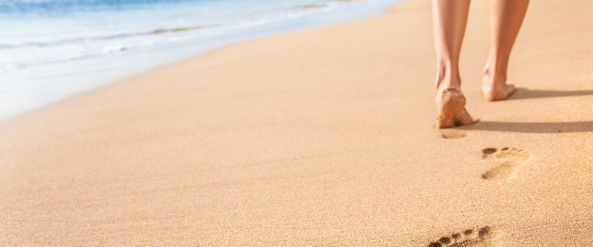 Pękające pięty - jak je wyleczyć? Zbliżenie na nogi kobiety i odciski stóp na piasku na plaży nad oceanem.