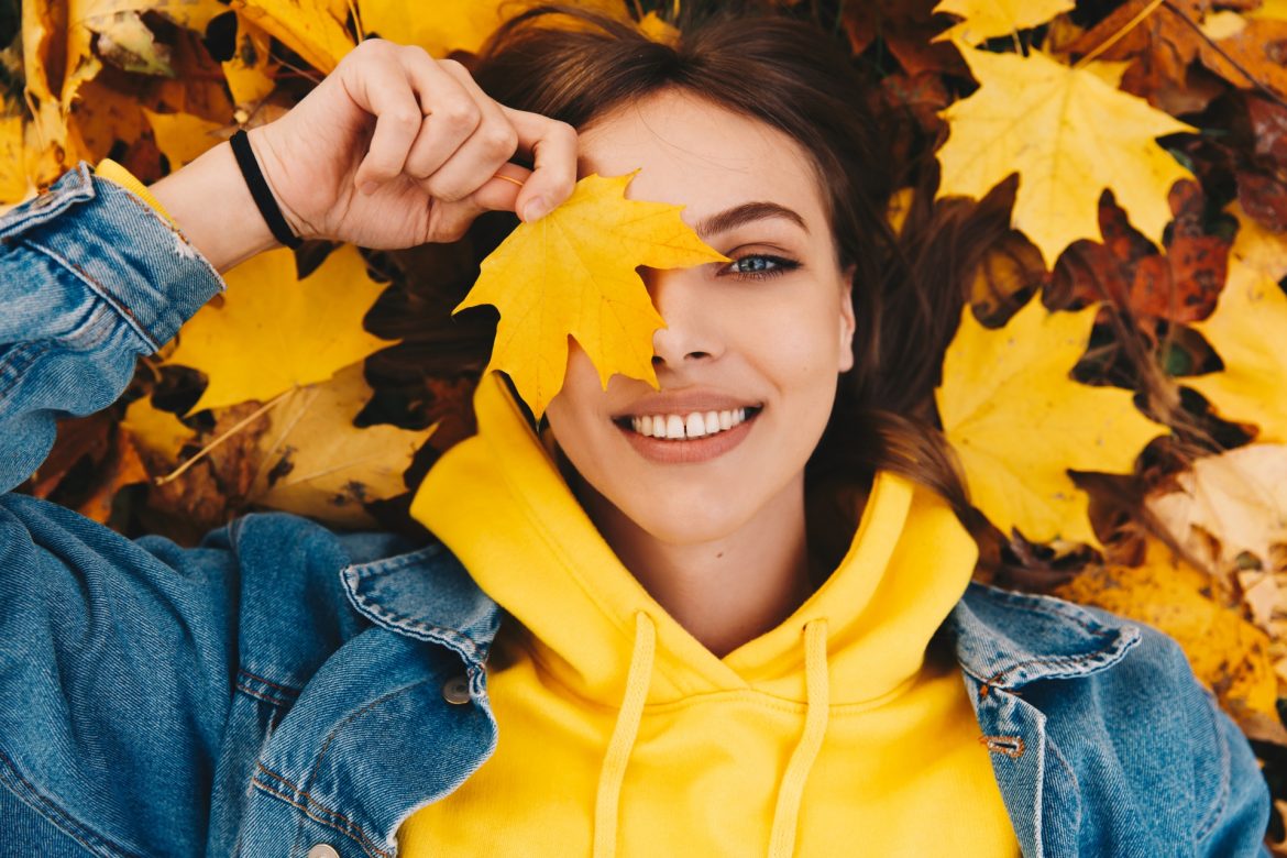 Odporność - czym jest, jak działa i jak ją budować? Dziewczyna w żółtej bluzie i kurtce jeansowej leży w jesiennych liściach i trzyma żółty liść przy twarzy, zasłaniając nim oko.
