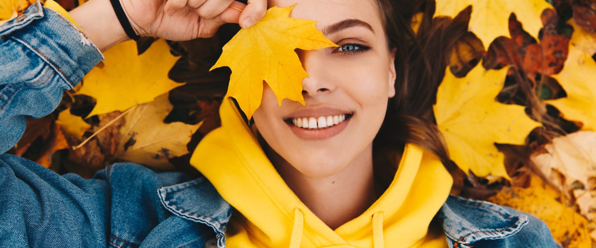 Odporność - czym jest, jak działa i jak ją budować? Dziewczyna w żółtej bluzie i kurtce jeansowej leży w jesiennych liściach i trzyma żółty liść przy twarzy, zasłaniając nim oko.