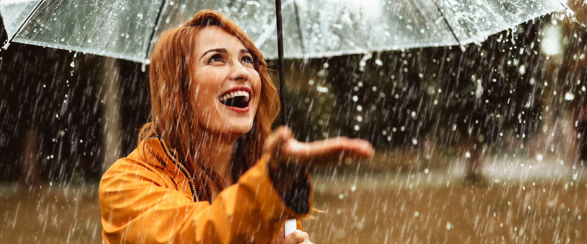 Czynniki obniżające odporność. Co wpływa na obniżoną odporność? Kobieta w żółtym płaszczyku pod przezroczystym parasolem wystawia rękę na deszcz.