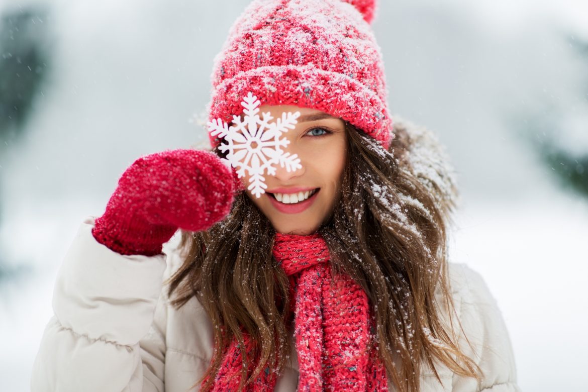 Jak wzmocnić odporność w 7 krokach? Kobieta w kurtce puchowej, czerwonej czapce i szaliku w zimowej scenerii uśmiecha się do aparatu, przykładając do oka ozdobny płatek śniegu.