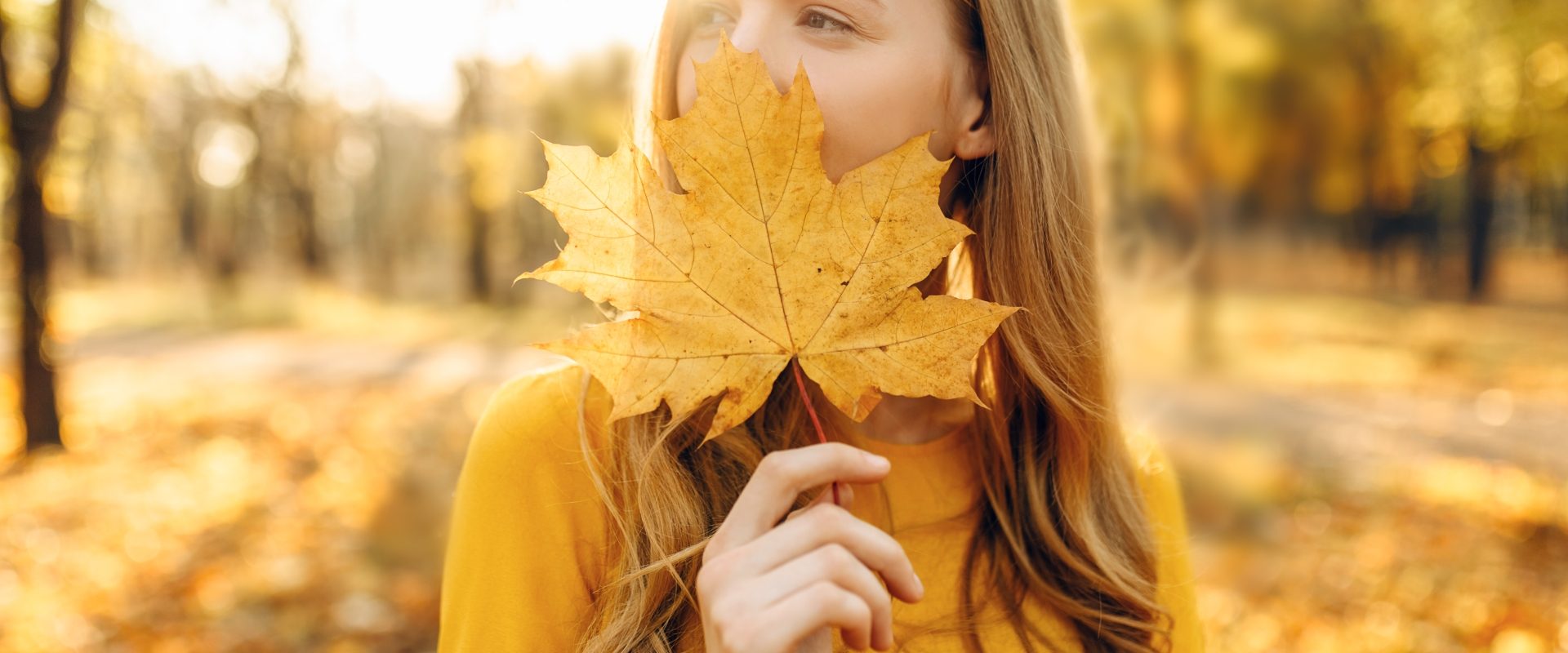 Naturalne sposoby na wzmocnienie odporności. Jak wzmocnić odporność jesienią i zimą? Młoda dziewczyna stoi w lesie i przykłada jesienny żółty liść do twarzy.