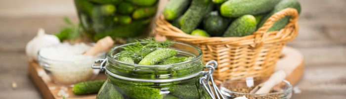 Przepis na ogórki kiszone i ogórki małosolne - jak zrobić ogórki kiszone i małosolne? Ogórki w słoikach i koszyczku wiklinowym na starym drewnianym stole.