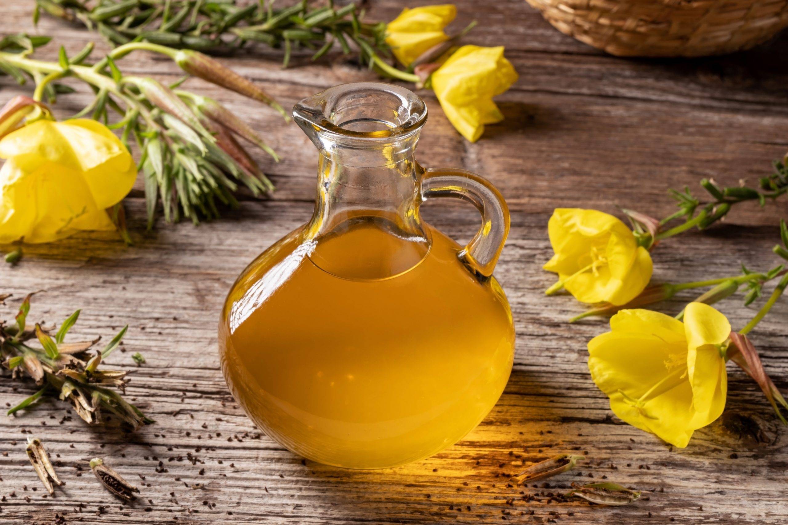 Olejek z wiesiołka - właściwości lecznicze i zastosowanie. Olej wiesiołkowy w szklanym dzbanku i żółte kwiaty wiesiołka na drewnianym stole.
