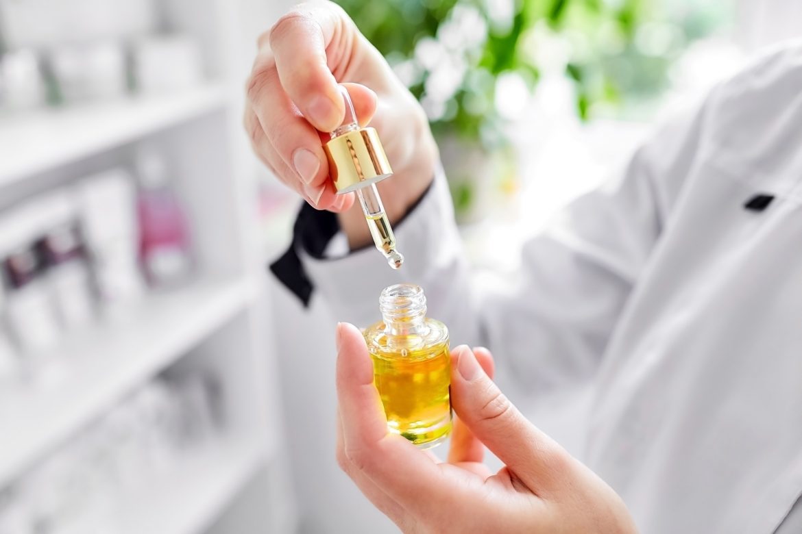 Olejek arganowy w pielęgnacji. Farmaceutka lub kosmetolog trzyma w ręku otwartą buteleczkę z olejkiem i podnosi pipetę do góry.