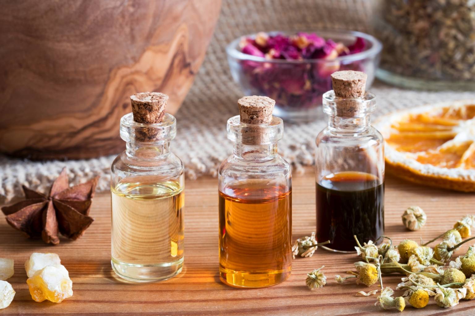 Jak działa aromaterapia? Czy olejki eteryczne mają właściwości lecznicze? Trzy buteleczki z olejkami eterycznymi stoją na drewnianym blacie w otoczeniu suszonych kwiatów, przypraw i cytrusów.