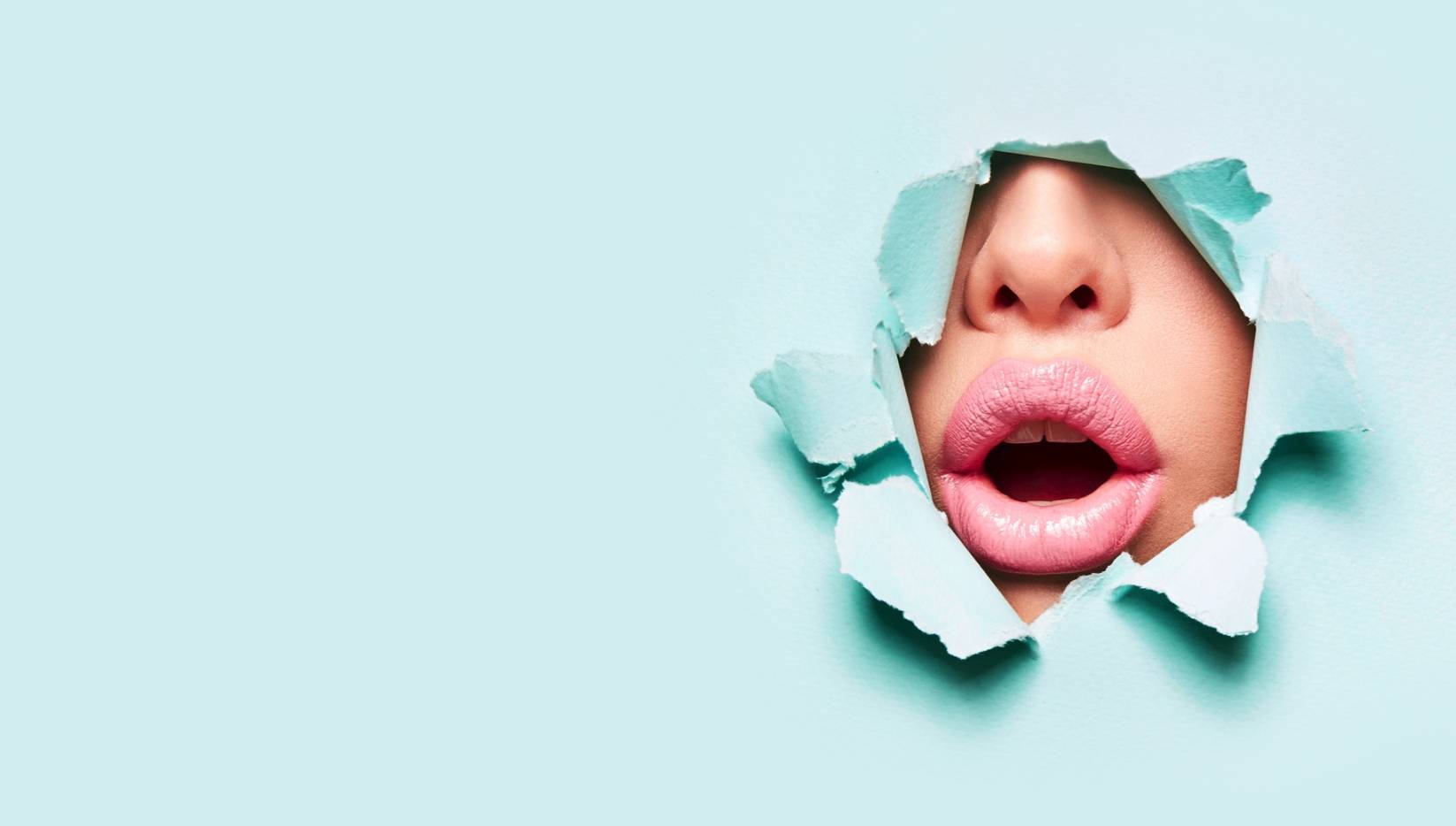 Opryszczka na ustach - czym jest, jak można się nią zarazić i jak z nią walczyć? Zdjęcie studyjne - kobieta o pełnych różowych ustach przebija się twarzą przez błękitny papier robiący za tło.