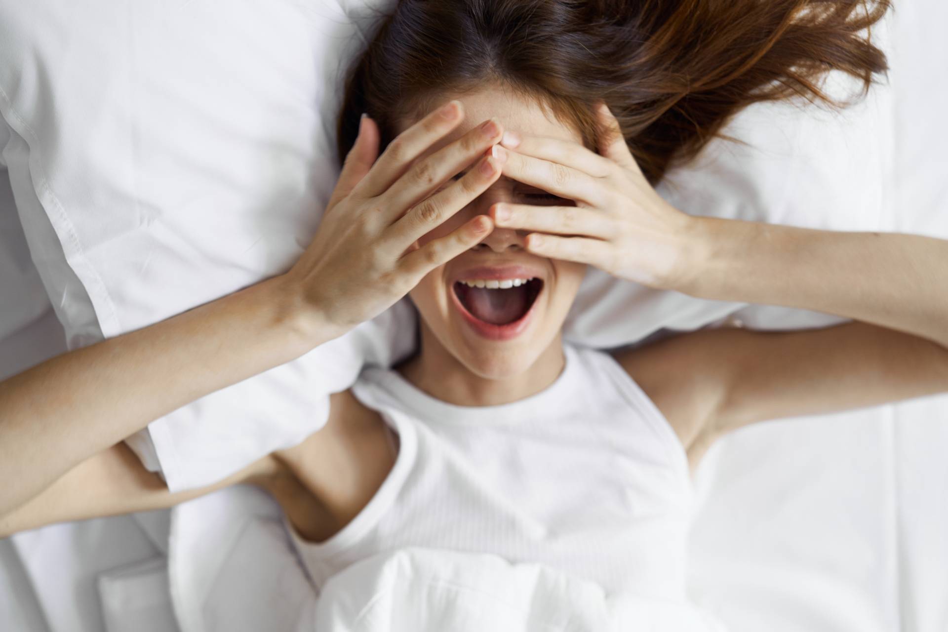 Masturbacja kobiet - jak się masturbować? Kobieta leży na łóżku i zasłania dłońmi oczy.