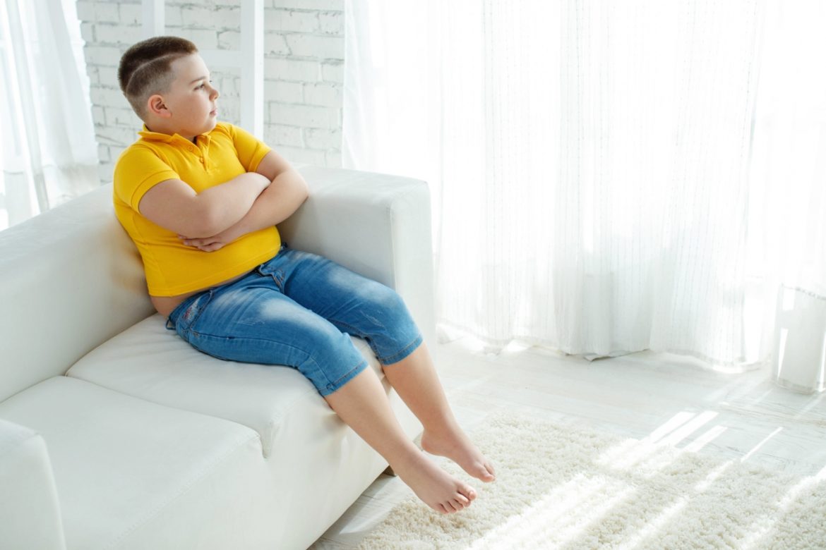 Otyłość u dzieci - jakie są jej przyczyny, skutki i jak ją leczyć? Otyły chłopiec siedzi na kanapie i patrzy w okno.