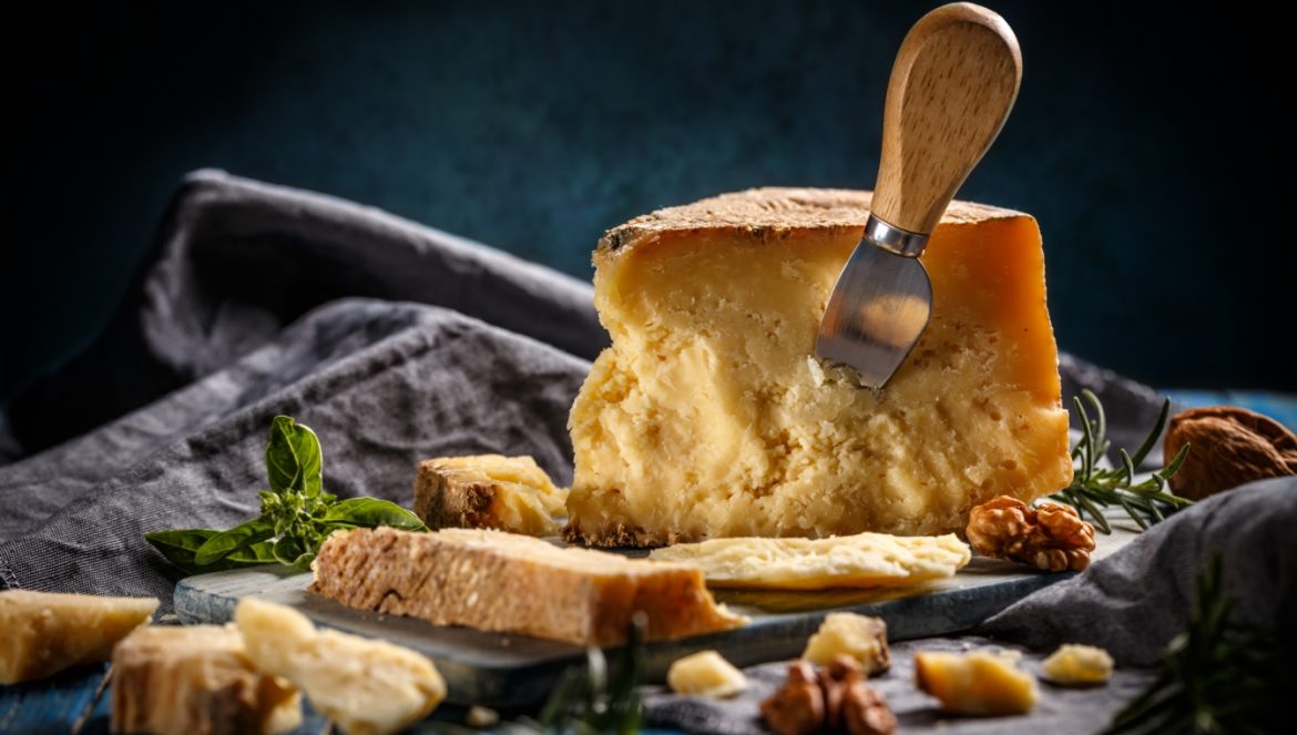 Parmezan - włoski król serów. Jakie właściwości odżywcze ma parmezan? Do czego go podawać? Sprawdź przepisy z parmezanem Agnieszki Żelazko. Kawałek sera parmezanu z wbitym nożem do sera na szarej tkaninie na czarnym tle.